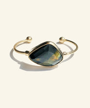 Load image into Gallery viewer, This faceted blue tigereye sits gleaming in a golden encasement. A shimmering dark blue with streaks of gold, makes this stone mysterious and sophisticated. The classic setting and golden colour displays the stone perfectly. The two orbs at the ends adds to comfort.