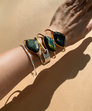 Load image into Gallery viewer, A pack of gleaming tigress bangles shown on an arm with the Pigna Arrow bangle classic. The faceted blue tigereyes sits in their golden encasements. Shimmering dark blue with streaks of gold, tigereye is a stone full of mystery and sophistication. The classic setting and golden colour displays the stone perfectly. The two orbs at the ends adds to comfort. 

