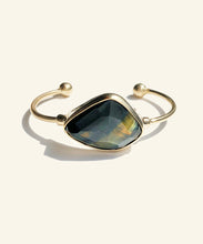 Load image into Gallery viewer, This faceted blue tigereye sits gleaming in a golden encasement. A shimmering dark blue with streaks of gold, makes this stone mysterious and sophisticated. The classic setting and golden colour displays the stone perfectly. The two orbs at the ends adds to comfort. 
