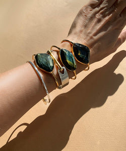 A pack of gleaming tigress bangles shown on an arm with the Pigna Arrow bangle classic. The faceted blue tigereyes sits in their golden encasements. Shimmering dark blue with streaks of gold, tigereye is a stone full of mystery and sophistication. The classic setting and golden colour displays the stone perfectly. The two orbs at the ends adds to comfort. 